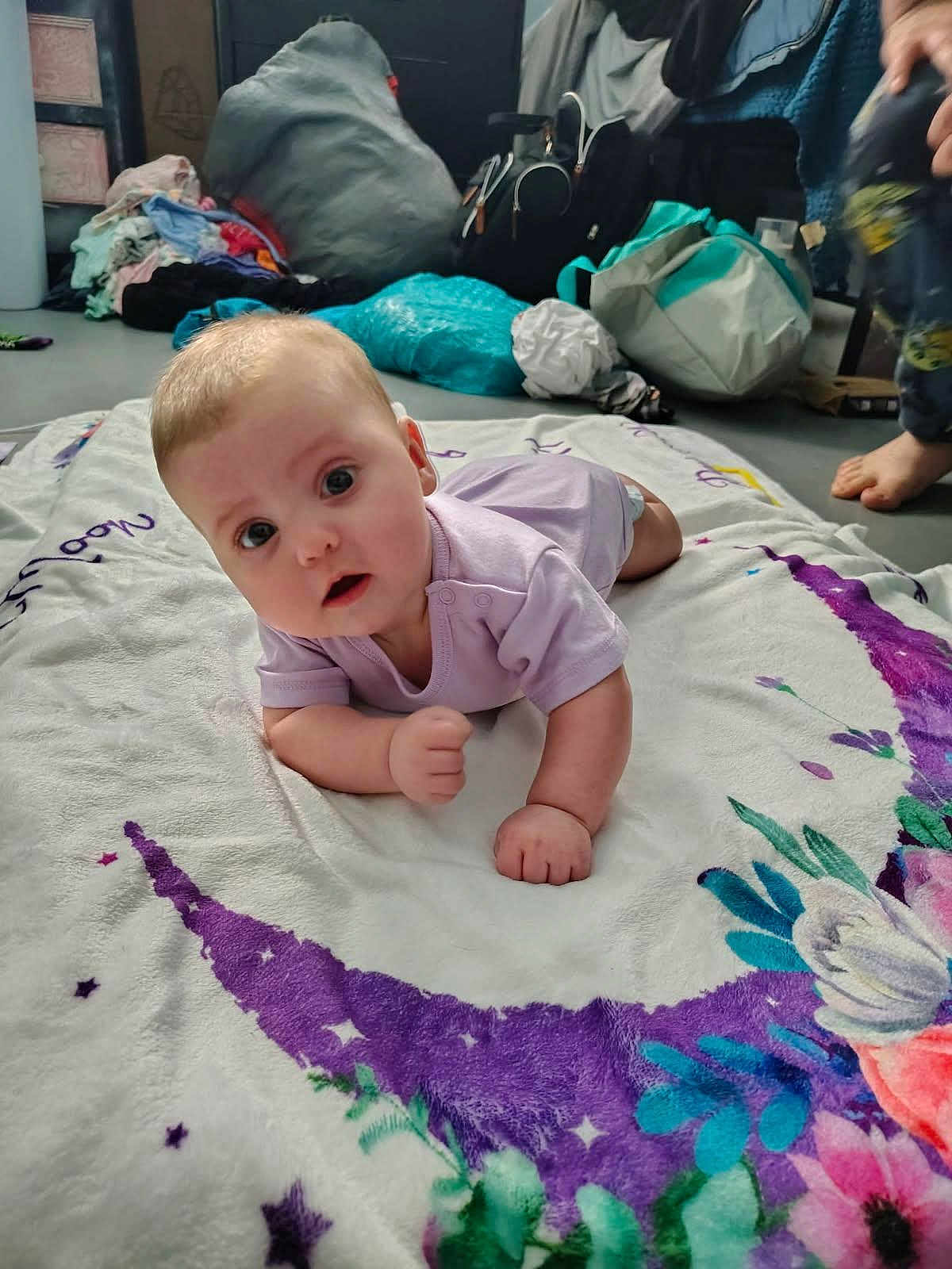 Morgna joined the competition — help win amazing prizes! baby, child, infant, blanket, purple, crescent_moon, floral, floor, indoors, clothing, onesie, curious, face, expression, hand, foot, person, background, bag, pile_of_clothes