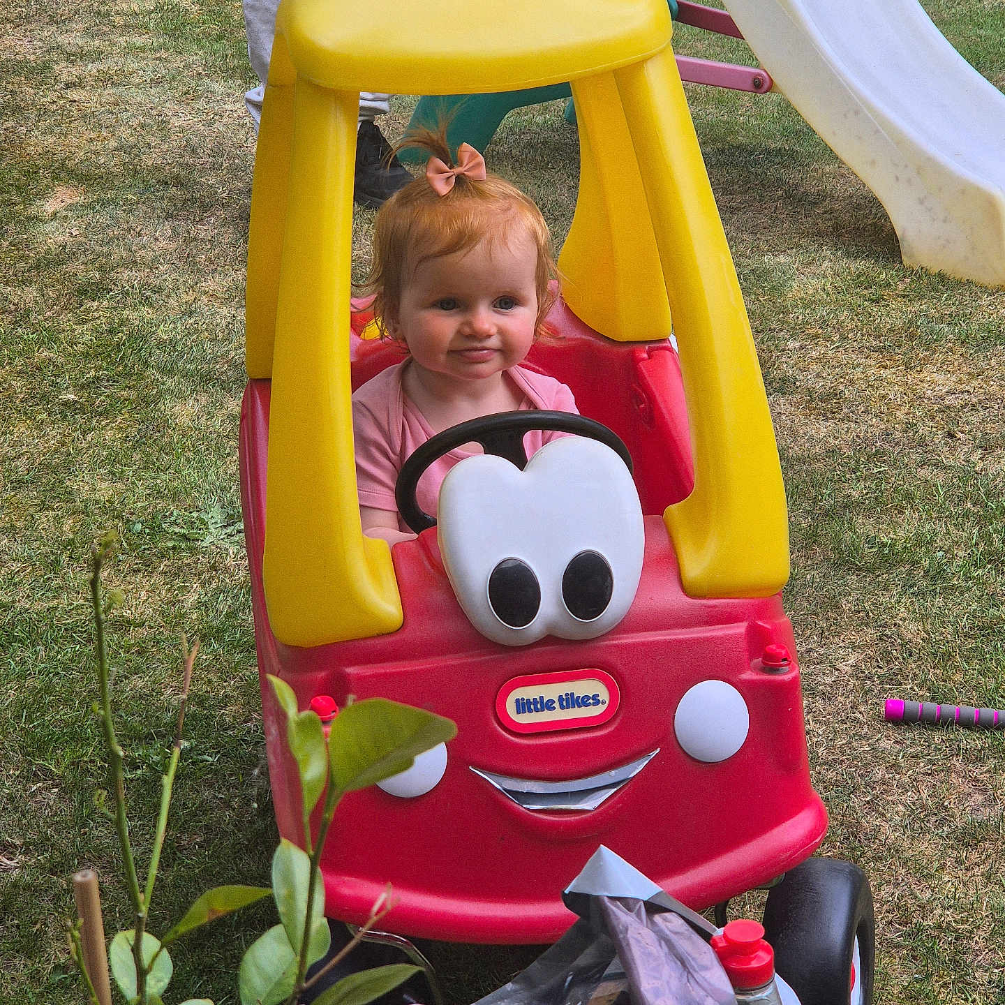 Keeley is registered to the contest to win money with this photo: adult, backyard, bow, child, grass, greenery, happy, outdoor, person, pink_clothing, plastic_toy, play, playground, red_car, slide, smile, steering_wheel, toddler, toy_car, yellow_roof