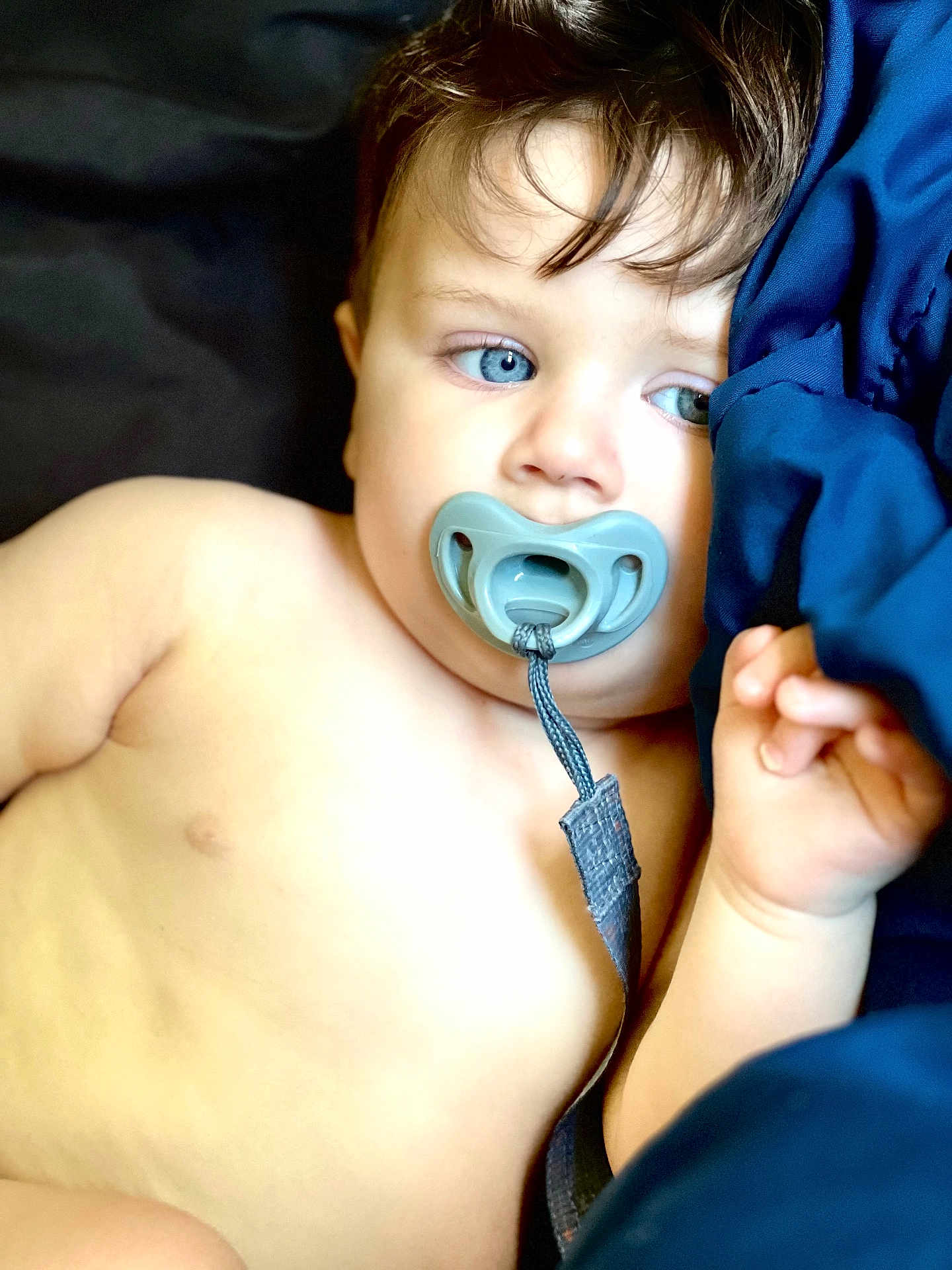Cash is registered to the contest to win money with this photo: baby, child, pacifier, blue_eyes, close_up, infant, skin, hand, portrait, lying_down, blue_fabric, straps, cute, young, face, expression, indoors, soft_light, resting, sleepy