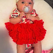 Emilia is registered to the contest to win money with this photo: baby, pacifier, headband, red_skirt, sandals, blanket, cushion, infant, clothing, footwear, person, indoors, cute, portrait, child, baby_girl, sitting, soft_toy, fabric, accessory
