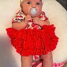 baby, pacifier, headband, red_skirt, sandals, blanket, cushion, infant, clothing, footwear, person, indoors, cute, portrait, child, baby_girl, sitting, soft_toy, fabric, accessory