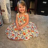 child, girl, smile, dress, floral_pattern, carpet, indoor, furniture, mattress, plastic_wrap, happy, portrait, sitting, short_hair, person, room, cabinet, clothing, floor, casual