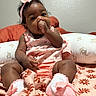 baby, infant, child, thumb_sucking, pink_clothing, headband, sock, blanket, floral_pattern, pillow, teddy_bear, cute, indoors, portrait, sitting, curly_hair, soft_lighting, young_child, comfort, adorable