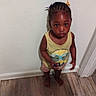 child, toddler, barefoot, braided_hair, colorful_clips, yellow_tank_top, printed_clothing, shorts, wooden_floor, indoor, white_wall, curious_expression, standing, person, casual_clothing, home, young_child, portrait, looking_up, floor