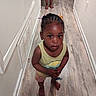 barefoot, braided_hair, casual_clothing, child, close_up, colorful_clips, curious, feet, hallway, home, indoors, looking_up, person, portrait, small_object, standing, toddler, white_wall, wooden_floor, young_child