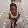 toddler, child, hallway, barefoot, floor, wooden_floor, indoor, person, standing, curious, yellow_shirt, shorts, braided_hair, hair_accessories, skin, hands, feet, adult_feet, wall, home