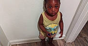 Kalani Douglas joined the competition — help win amazing prizes! child, toddler, indoor, barefoot, yellow_tank_top, minion_graphic, shorts, wooden_floor, corner, curly_hair, braids, wall, door_frame, looking_up, standing, person, skin, clothing, expression, flooring
