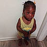toddler, child, indoor, barefoot, toy, yellow_shirt, shorts, wooden_floor, doorframe, wall, curly_hair, braids, standing, looking_up, expression, person, young_child, floor, holding, casual_clothing