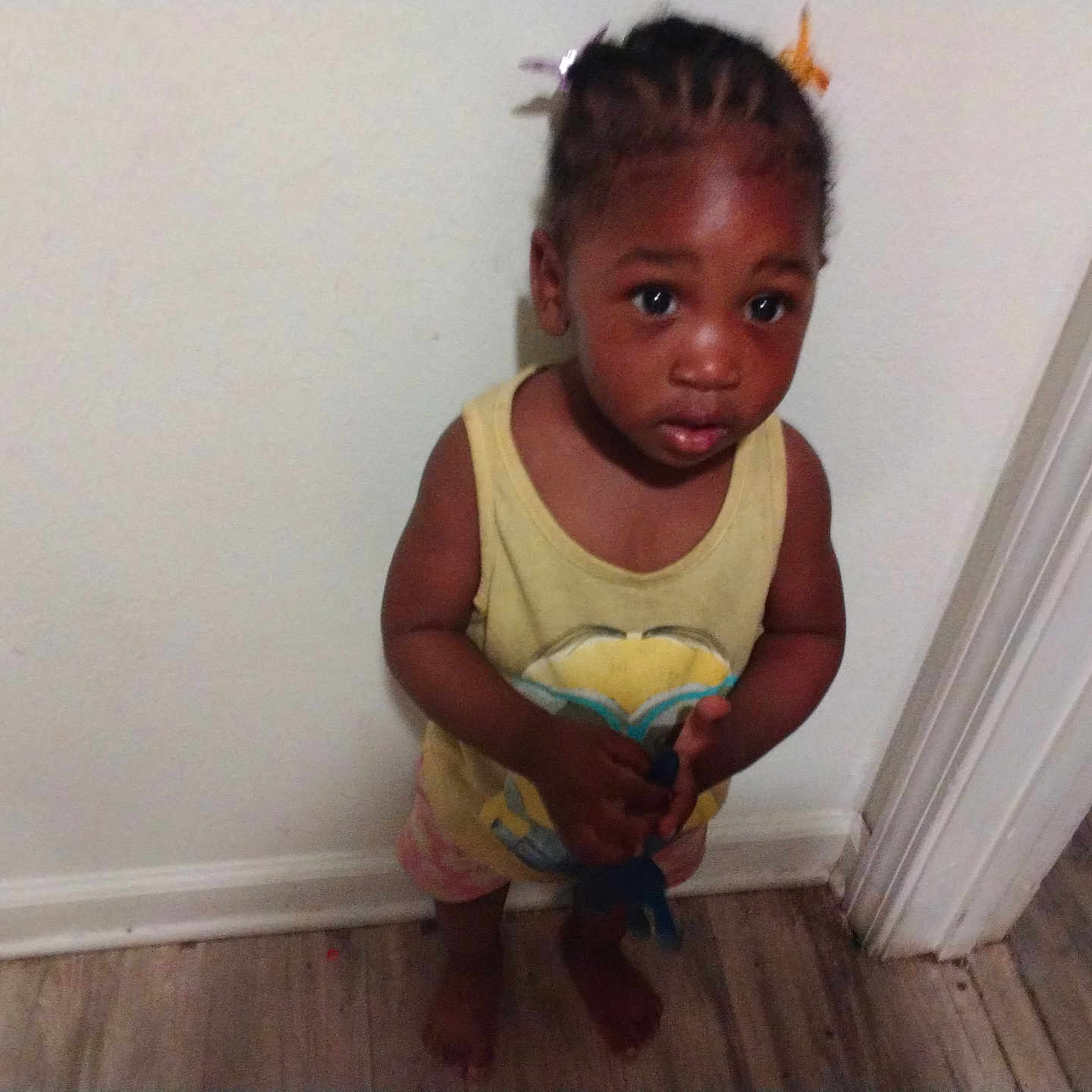 Kalani Douglas is registered to the contest to win money with this photo: toddler, child, indoors, barefoot, braided_hair, yellow_shirt, shorts, wooden_floor, white_wall, doorframe, holding_object, curious, standing, young_child, person, portrait, expression, colorful_clips, home, flooring