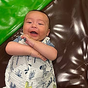 Gabriel a rejoint le concours — aidez-le/la à gagner de superbes lots ! animal_print, baby, brown_surface, child, closeup, crossed_arms, cute, expression, green_cushion, happy, indoors, infant, lying_down, person, playful, portrait, skin, smiling, white_outfit, young_child