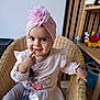 baby, child, girl, pink_turban, flower, wicker_chair, smile, finger_on_mouth, long_sleeve_shirt, leggings, toy, shelf, wooden_shelf, indoor, cute, playful, face, person, furniture, happy