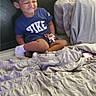 child, toddler, bed, blanket, bedroom, sitting, cross_legged, scrunching_face, smile, toy, shirt, nike, socks, pillows, bedding, indoor, portrait, casual, phone_screen, gallery_interface
