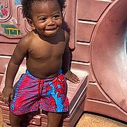 King joined the competition — help win amazing prizes! child, toddler, smiling, shirtless, curly_hair, earrings, swim_trunks, spiderman, crocs, playground, slide, outdoor, sunlight, happy, sitting, concrete, plastic, play_area, summer, cute