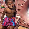 child, toddler, smiling, shirtless, curly_hair, earrings, swim_trunks, spiderman, crocs, playground, slide, outdoor, sunlight, happy, sitting, concrete, plastic, play_area, summer, cute