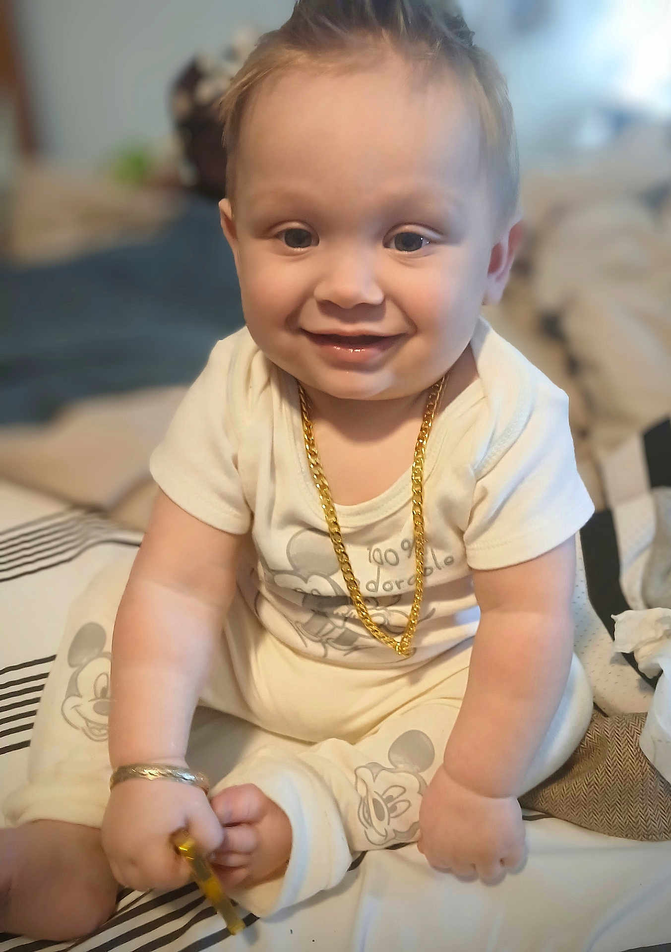 Anthony is registered to the contest to win money with this photo: baby, child, smiling, jewelry, gold_chain, bracelet, mickey_mouse, pajamas, sitting, bed, indoor, cute, happy, infant, person, face, hand, foot, clothing, toy