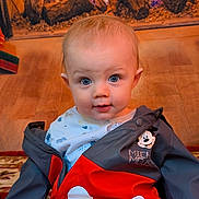 Brodie is registered to the contest to win money with this photo: baby, blue_eyes, child, clothing, cozy, curious, cute, expression, fireplace, floor_rug, home, indoor, infant, jacket, mickey_mouse, portrait, sitting, smile, warm, wood_floor