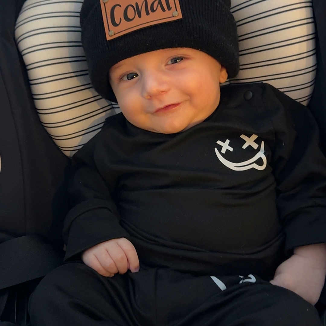 Conal is registered to the contest to win money with this photo: apparel, baby, black_clothing, child, cozy, cute, face, footwear, hands, happy, hat, headwear, indoors, infant, legs, person, sitting, smile, striped_cushion, young