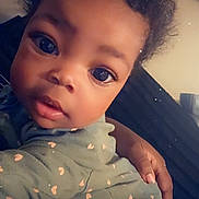 Briella is registered to the contest to win money with this photo: baby, infant, child, portrait, close_up, wide_eyes, looking_at_camera, curly_hair, heart_emoji, green_outfit, patterned_clothing, adult_hand, being_held, indoor, bed, bedding, cozy, face, photography, vertical_photo
