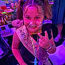 child, girl, party, cake, face_paint, sash, smile, peace_sign, glowing_necklaces, pigtails, celebration, indoor, happy, person, event, colorful, fun, people, table, food