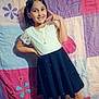 casual, child, colorful_background, cute, dress, fashion, floor, girl, happy, heart_icon, indoors, necklace, playful, portrait, pose, quilt, smiling, standing, white_shoes, young
