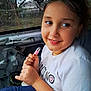 child, girl, smile, car_interior, white_tshirt, holding_object, pink, short_hair, jewelry, window, outdoor, trees, fence, seat, candid, portrait, happy, casual, daylight, young