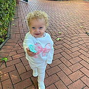 Marley Rose joined the competition — help win amazing prizes! child, toddler, curly_hair, blue_eyes, pacifier, white_clothing, sweatshirt, sneakers, brick_pathway, outdoor, greenery, sunlight, smiling, walking, person, playful, nature, daylight, casual_wear, cute
