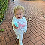 child, toddler, curly_hair, blue_eyes, pacifier, white_clothing, sweatshirt, sneakers, brick_pathway, outdoor, greenery, sunlight, smiling, walking, person, playful, nature, daylight, casual_wear, cute