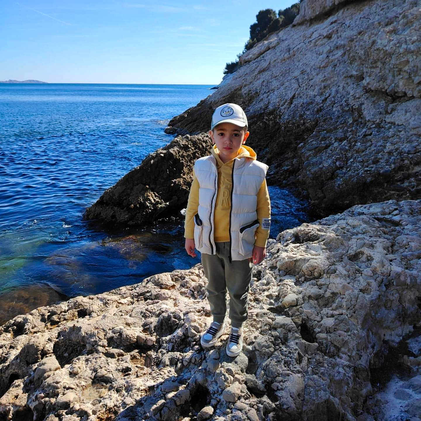 Aylan participe au concours pour gagner de l'argent avec cette photo : boy, cap, casual_clothing, child, coast, daytime, hoodie, nature, outdoor, pants, portrait, rocky_shore, scenery, sea, shoes, sky, standing, sunlight, vest, water