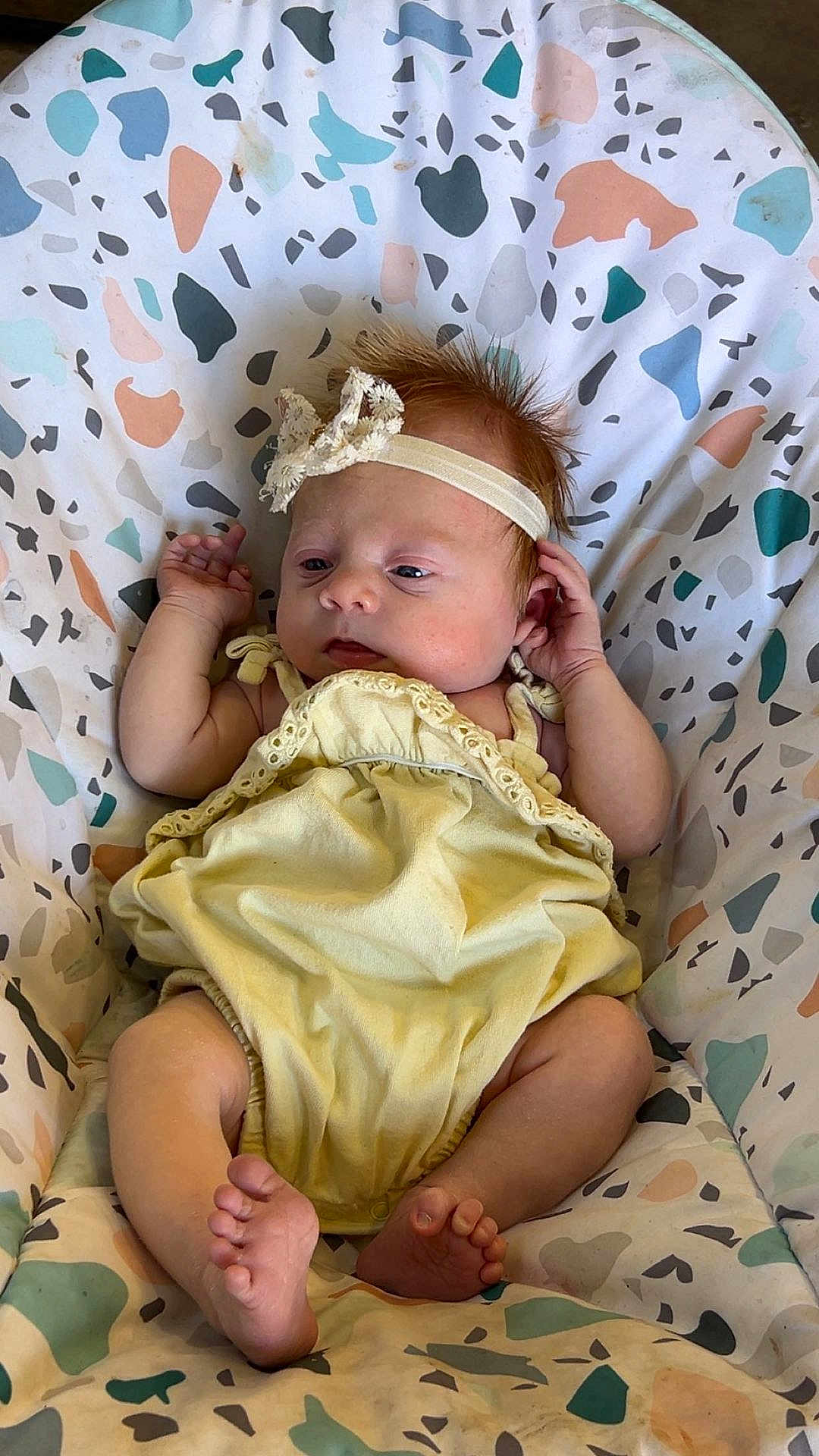 Maggie joined the competition — help win amazing prizes! baby, infant, child, yellow_outfit, headband, floral_headband, patterned_seat, colorful_background, barefoot, small_hands, small_feet, relaxed, indoors, portrait, cute, young, sitting, comfortable, soft_lighting, skin