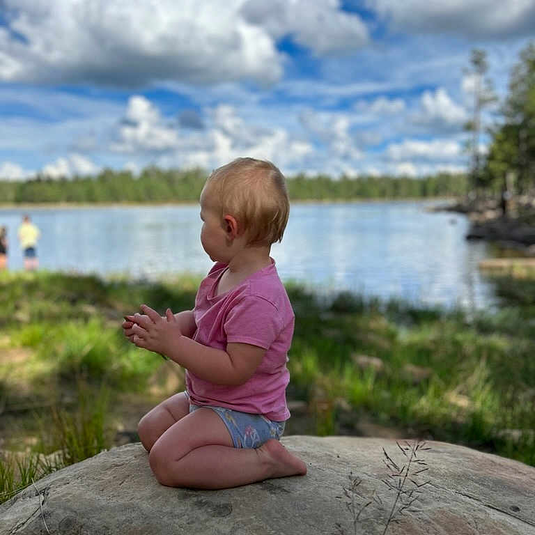 Everleigh joined the competition — help win amazing prizes! baby, clothing, cloud, face, grass, head, lake, nature, outdoors, person, photography, plant, portrait, rock, scenery, shorts, sitting, sky, water, waterfront