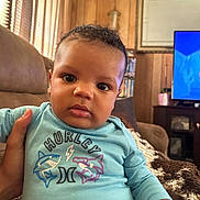 Damauirre is registered to the contest to win money with this photo: baby, child, indoor, person, blue_shirt, shark_graphic, couch, hand, wood_paneling, television, blanket, face, eyes, curly_hair, furniture, living_room, casual_clothing, expression, portrait, home