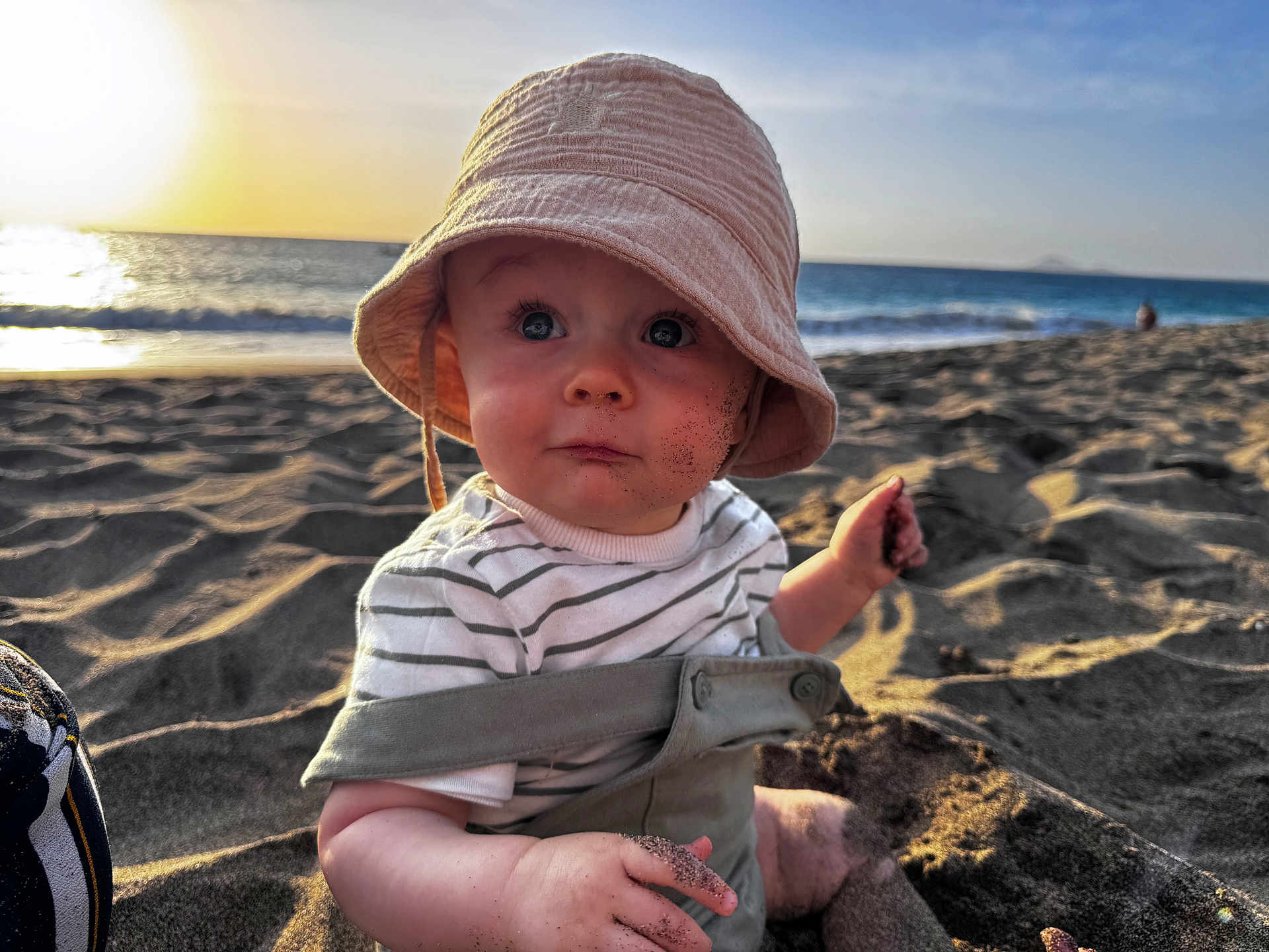 Hugo participe au concours pour gagner de l'argent avec cette photo : baby, child, beach, sand, sunset, hat, striped_shirt, ocean, water, waves, sitting, curious, face, hand, outdoor, sunlight, nature, play, vacation, portrait