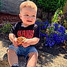 toddler, child, outdoor, flower, plant, rock, leaf, sunlight, shoes, shorts, tshirt, curly_hair, garden, greenery, nature, casual_clothing, sitting, daylight, happy, portrait