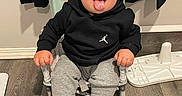 Xander joined the competition — help win amazing prizes! baby, child, tongue_out, sitting, black_hoodie, grey_sweatpants, sneakers, indoor, wooden_floor, chair, playful, cute, person, flooring, clothing, young_child, smiling, tongue, black_clothing, casual