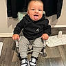 baby, child, tongue_out, sitting, black_hoodie, grey_sweatpants, sneakers, indoor, wooden_floor, chair, playful, cute, person, flooring, clothing, young_child, smiling, tongue, black_clothing, casual