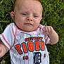 baby, infant, grass, outdoor, clothing, white_shirt, baseball_jersey, detroit_tigers, person, face, head, blue_eyes, hand, fist, greenery, nature, portrait, young_child, casual, summer