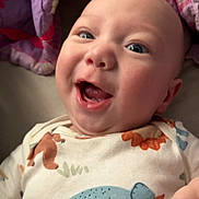 Asiyah is registered to the contest to win money with this photo: baby, infant, smiling, happy, face, dinosaur_onesie, clothing, blanket, purple, floral_pattern, cute, child, young, portrait, indoor, person, skin, head, hand, expression