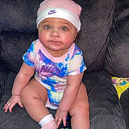 La Niyah is registered to the contest to win money with this photo: baby, child, infant, onesie, hat, nike, remote_control, couch, sofa, sitting, pink_socks, colorful_clothing, indoor, cute, chubby_cheeks, face, person, smiling, home, plush