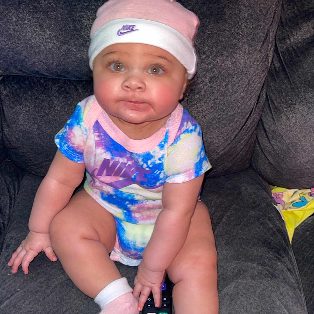 La Niyah is registered to the contest to win money with this photo: baby, child, infant, onesie, hat, nike, remote_control, couch, sofa, sitting, pink_socks, colorful_clothing, indoor, cute, chubby_cheeks, face, person, smiling, home, plush