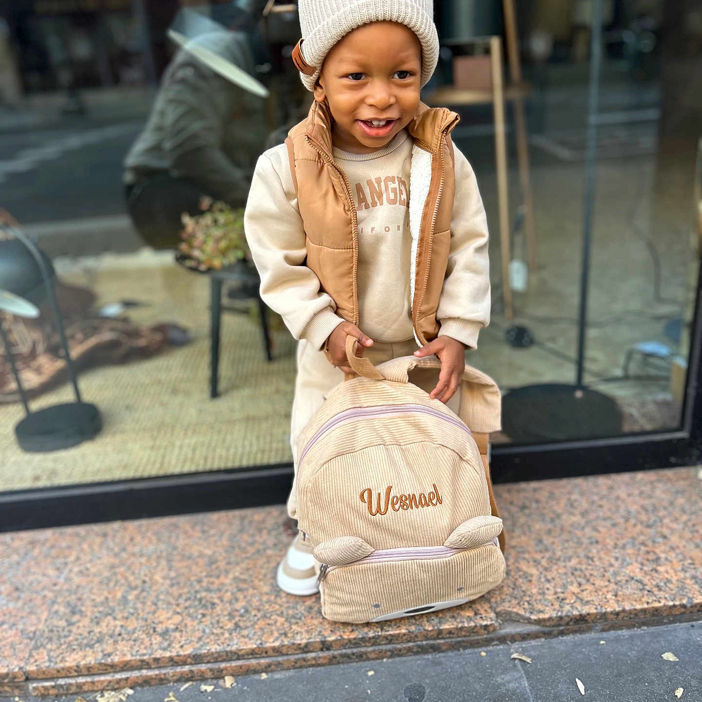 Wesna participe au concours pour gagner de l'argent avec cette photo : accessory, backpack, beanie, beige, candid, child, fashion, hands, portrait, pose, reflection, shoes, shop_window, sidewalk, smile, street, sweatshirt, toddler, urban, vest