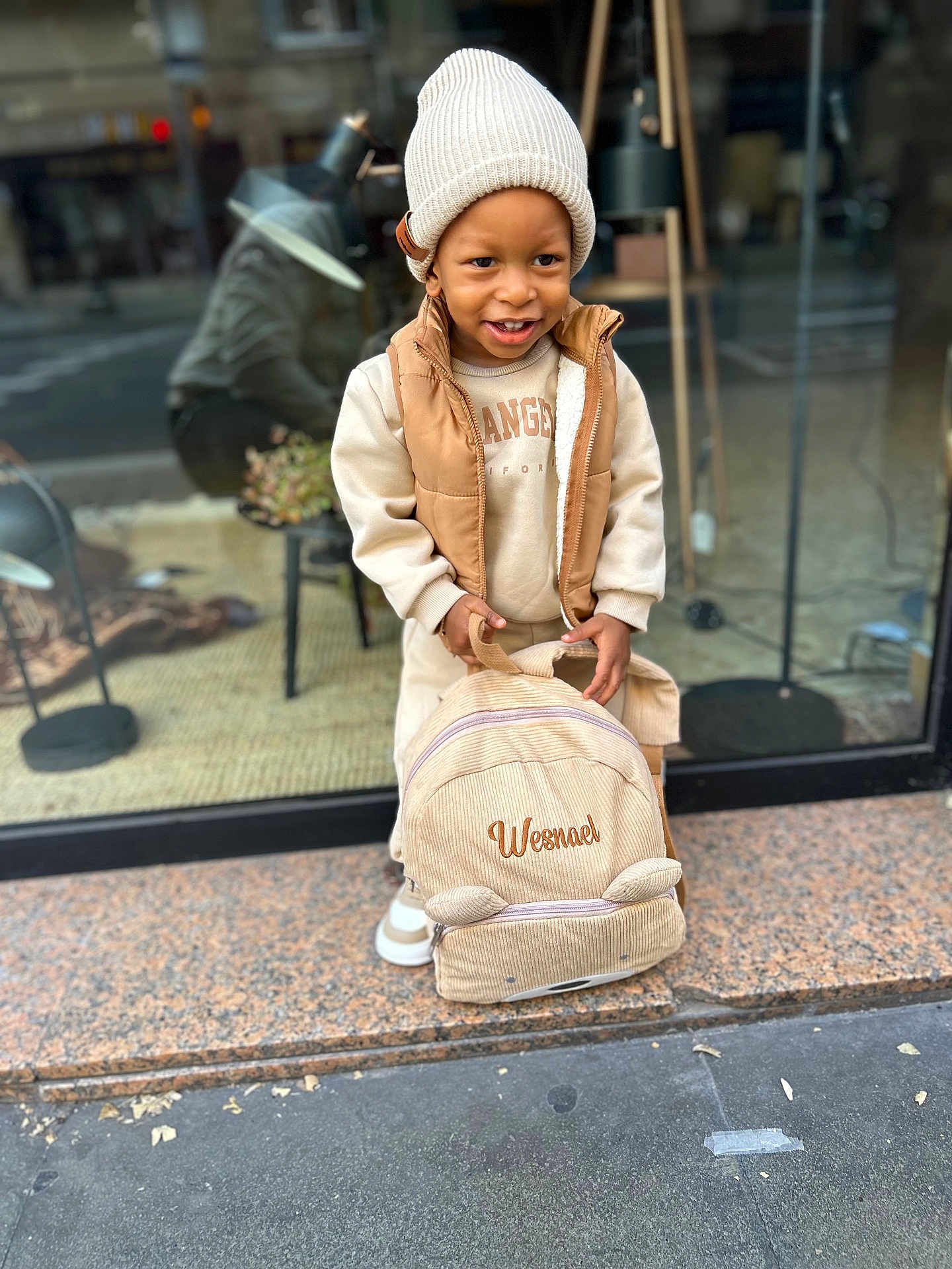 Wesna participe au concours pour gagner de l'argent avec cette photo : child, toddler, beanie, backpack, vest, sweatshirt, smile, street, sidewalk, shop_window, reflection, portrait, candid, urban, shoes, beige, fashion, accessory, hands, pose