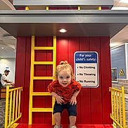 Zayaan joined the competition — help win amazing prizes! child, playground, indoor, red_hair, orange_shirt, yellow_ladder, safety_sign, crouching, play_structure, adult, bench, carpet, ceiling_lights, smiling, curly_hair, casual_clothing, play_area, rules, no_climbing, no_throwing