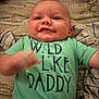 Blaine is registered to the contest to win money with this photo: baby, infant, smile, green_shirt, text_on_clothing, lying_down, blanket, onesie, happy, cute, hand_motion, face, child, clothing, fabric, casual, indoors, close_up, person, soft_texture