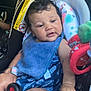 baby, child, car_seat, blue_bib, blue_outfit, toy, curly_hair, car_interior, seatbelt, infant, child_seat, relaxed, cute, hand, leg, colorful, fabric, seat, sitting, portrait