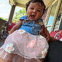 baby, blue_top, casual, child, curly_hair, daylight, dress, flower, happy, holding, legs, outdoor, person, pink, porch, railing, smiling, tricycle, tutu, window