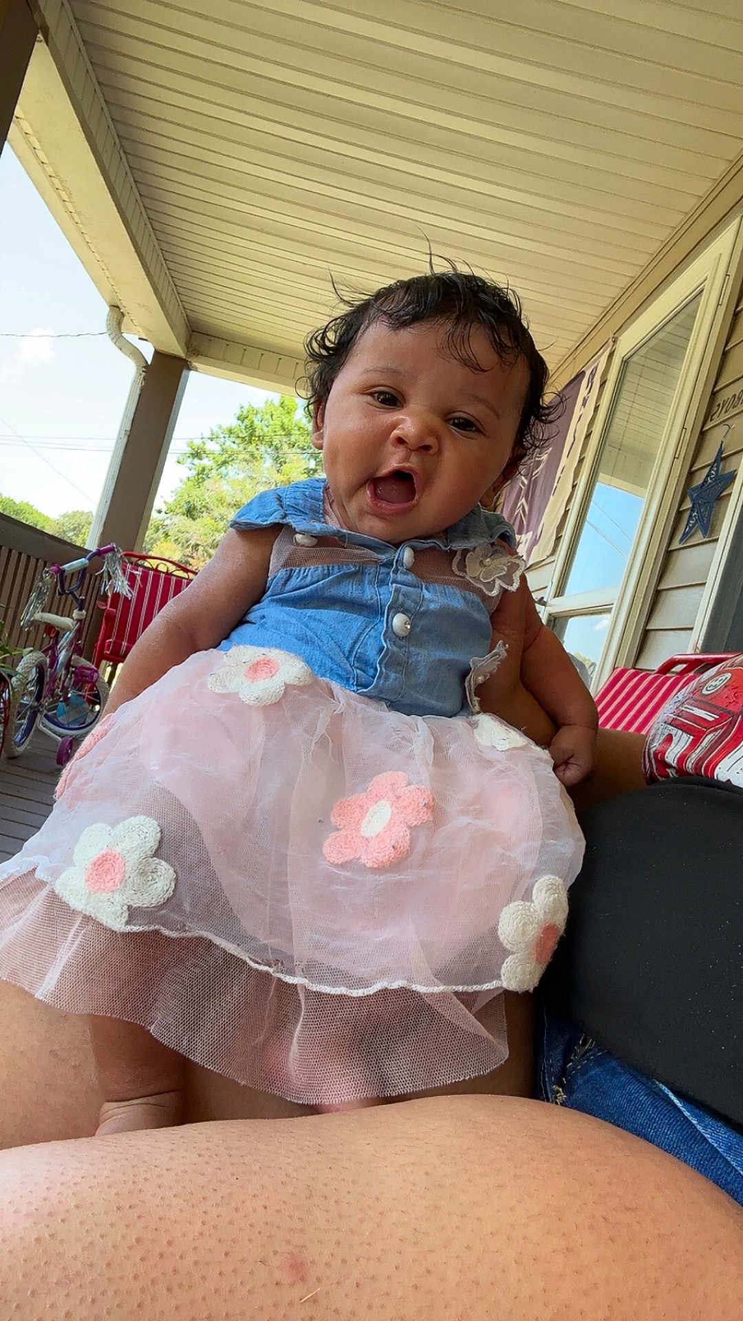 Harmoniee is registered to the contest to win money with this photo: baby, child, tutu, dress, blue_top, pink, flower, porch, outdoor, person, tricycle, curly_hair, smiling, happy, holding, legs, window, railing, daylight, casual
