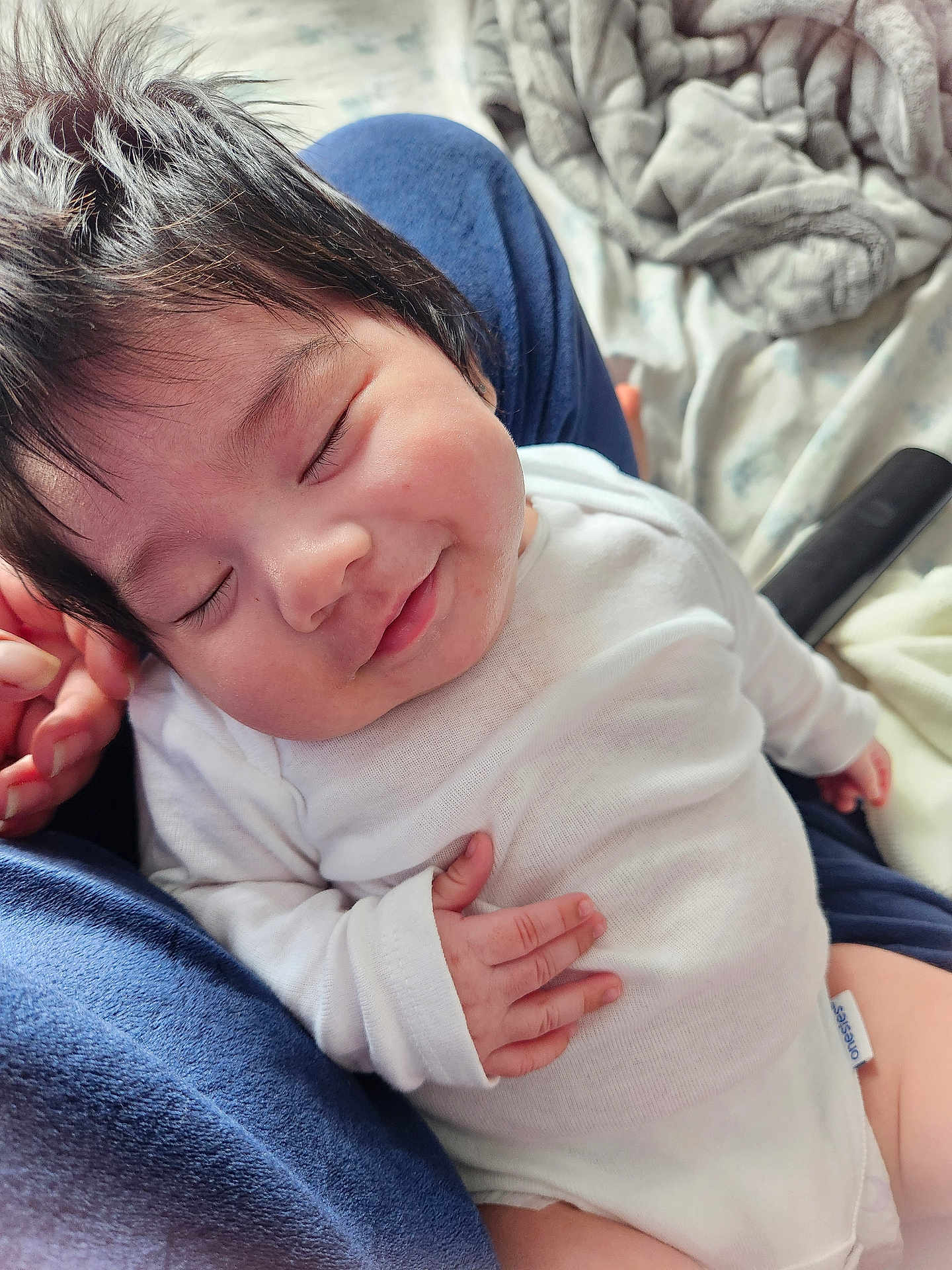 Nefertari joined the competition — help win amazing prizes! baby, infant, newborn, sleeping, smiling, white_onesie, hand_on_chest, tousled_hair, closeup, portrait, lap, blanket, bedding, cozy, soft_texture, indoor, cute, tiny_fingers, parent_hand, relaxed