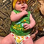 baby, child, closeup, clothing, cute, diaper, face, green_shirt, hands, happy, headband, indoors, infant, legs, patterned_blanket, person, sitting, smiling, toddler, yellow_bow