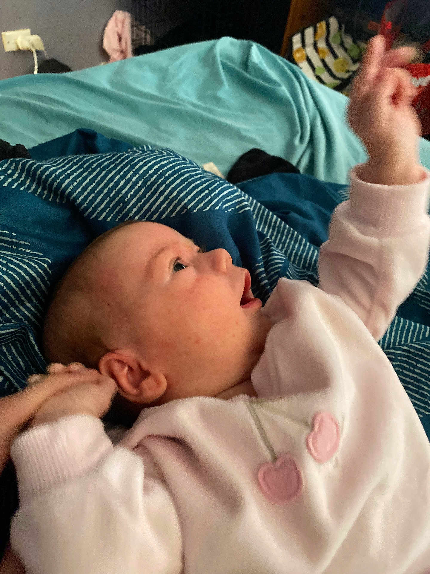 Aria is registered to the contest to win money with this photo: baby, infant, child, bed, blanket, blue_blanket, teal_blanket, pink_clothing, heart_design, hand, face, ear, skin, lying_down, indoor, soft_fabric, cute, young_child, expression, reaching_hand