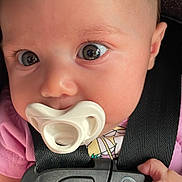 Lisboa is registered to the contest to win money with this photo: baby, infant, pacifier, car_seat, child, face, hand, clothing, pink, closeup, young_child, sitting, seatbelt, cute, curious, portrait, indoor, human, person, newborn
